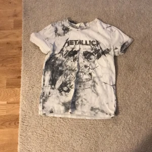 Vit och svart Metallica t-shirt - Säljer en cool vit t-shirt med Metallica-tryck och svart batikmönster. T-shirten har korta ärmar och ett stort tryck av en dödskalle på framsidan. Perfekt för rockälskare! 🤘