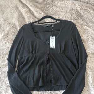Svart långärmad topp från Vero Moda - Svart långärmad topp från Vero Moda. Toppen har en söt rosett framtill och spetsdetaljer längs kanterna. Perfekt för en elegant look. HELT NY med prislapp, storlek m💕org. pris 349 kr