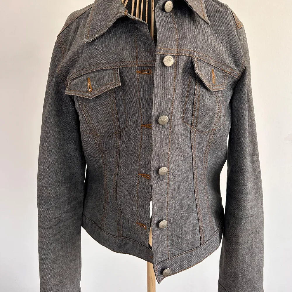 Snygg gråblå jeansjacka från Vero Moda med klassisk krage och knappar framtill. Jackan har två bröstfickor med knappdetaljer och kontrastsömmar i orange. Perfekt för en stilren look!. Takit.
