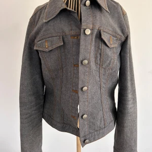 Gråblå jeansjacka från Vero Moda - Snygg gråblå jeansjacka från Vero Moda med klassisk krage och knappar framtill. Jackan har två bröstfickor med knappdetaljer och kontrastsömmar i orange. Perfekt för en stilren look!