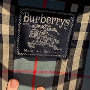 Blå trenchcoat från Burberry - Säljer en klassisk blå trenchcoat från Burberry av den äldre modellen med det ikoniska rutiga fodret. Jackan har knappar framtill och en stilren krage. Perfekt för en elegant look. Säljs pga. Graviditet. Storlek s/m