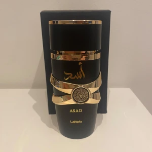 Asad Parfym från Lattafa - Säljer en elegant parfym vid namn Asad från Lattafa. Flaskan är svart med gyllene detaljer och har en lyxig design. Parfymen kommer i en stilren svart förpackning med dekorativa mönster. Perfekt för den som söker en sofistikerad doftupplevelse. Helt full