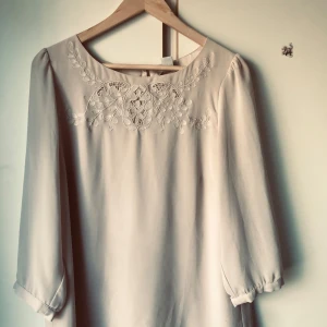 Beige blus med broderad detalj - Elegant beige klänning med vackert broderi på framsidan. Den har trekvartsärmar och en enkel, stilren design. Perfekt för en sofistikerad look.