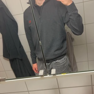 Mörkgrå Hugo Boss tröja med zip - Säljer en stilren mörkgrå tröja med dragkedja vid halsen. Tröjan har långa ärmar och en liten röd detalj på bröstet. Perfekt för en avslappnad look. Skick 9/10-10/10 Inga tecken på användning 