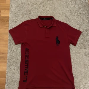 Röd pikétröja från Ralph Lauren - Säljer en snygg unik röd pikétröja från Ralph Lauren med klassisk krage och knappar. Tröjan har ett stort svart polospelarbroderi på framsidan och texten 'R.L. Challenge Cup' på sidan. Baksidan pryds av en polospelare och texten 'Challenge Cup MCMXLVII'. Denna piké säljs inte längre därav priset. I bra skick tvättad några fåtal gånger. 100 % ÄKTA