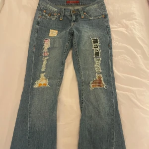 Blå jeans med patchwork och slitningar - Unika blå jeans med coola slitningar och färgglada patchwork-detaljer. De har en klassisk femficksdesign och knappar fram. Bakfickorna har snygga broderier som ger extra stil. Perfekta för en avslappnad look med en twist. De är väldigt lågmidjade och de e bootcut. Midjemått 38 innerbenslängd 72