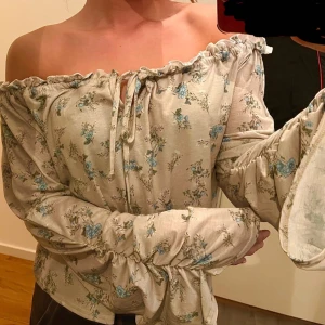 Romantisk offshoulder blus - Säljer en söt offshoulder blus med blommigt mönster i blått och beige. Blusen har långa ärmar med knytband och volangdetaljer vid ärmslut och halslinning. Perfekt för en romantisk look!
