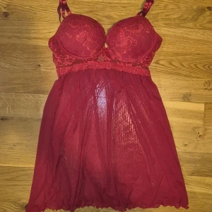 Röd spets lingerie  - Säljer en elegant röd spets lingerie/BH med vadderade kupor och justerbara axelband. Klänningen har en vacker spetsdetalj upptill och ett smått genomskinligt tyg nedtill som ger en romantisk känsla. Den är i princip oanvänd då jag inte brukar använda lingerie och det var en present. BH storleken är 75A, på bilderna ser den mer kallare ut i färgen men den har en varmare röd färg i verkligheten.