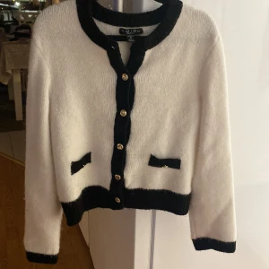 Vit och svart kofta från Misu - Snygg vit kofta med svarta detaljer från Misu. Den har silverknappar framtill och svarta kanter vid ärmslut och nederkant. Två dekorativa fickor framtill ger en chic touch. Perfekt för att lyfta din outfit!