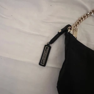 Svart axelväska med kedja från Victoria secret - Snygg svart axelväska från Victoria's Secret med en elegant guldkedja som axelrem. Väskan har en dragkedja upptill och en stilren design med en liten logotyp framtill. Perfekt för att ge en touch av glamour till din outfit.