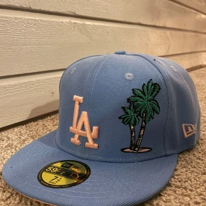 Blå keps med palm broderi från New Era - Snygg blå keps från New Era med broderade palmer och LA-logga framtill. Kepsen har en platt skärm och är en del av 59FIFTY-serien. Perfekt nu inför sommaren för att ge din stil en cool touch. Storlek: 7 1/2 (59.6 cm). Den är aldrig använd. Priset är diskuterbart. Skriv vid minsta fundering!