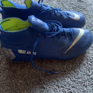 Blå Nike fotbollsskor - Säljer ett par blå Nike fotbollsskor med vita detaljer och snörning. Skorna har en hög ankelkrage och är designade för optimal prestanda på planen. Perfekta för fotbollsspelare som vill ha stil och funktionalitet.