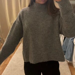 Grå stickad tröja - Säljer en mysig grå stickad tröja med hög krage/polo. Tröjan har en avslappnad passform och långa ärmar, perfekt för kyliga dagar. Den är enkel att matcha med olika outfits och ger en stilren look.