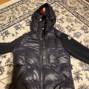 Svart pufferjacka från Moncler - Snygg svart pufferjacka från Moncler med huva och dragkedja framtill. Jackan har långa ärmar och flera praktiska fickor med dragkedjor. Perfekt för kyligare dagar.