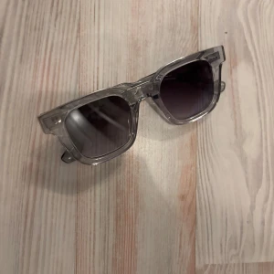 Chimi 04 - Snygga solglasögon med transparent båge och gråtonade linser. Perfekta för en stilren look. Bågarna har en klassisk fyrkantig form som passar de flesta ansikten.