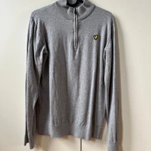 Grå tröja med dragkedja från Lyle & Scott - Säljer en stilren grå tröja från Lyle & Scott med dragkedja och hög krage. Tröjan har ett broderat emblem på bröstet och är tillverkad i ett mjukt material. Perfekt för en avslappnad look.