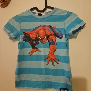 Blå randig Spiderman t-shirt från H&M - Cool blå randig t-shirt från H&M med ett stort Spiderman-tryck på framsidan. Perfekt för små superhjältefans! Tillverkad i mjuk bomull för en bekväm känsla hela dagen.