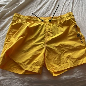 Gula badshorts från Sail Racing - Snygga gula badshorts från Sail Racing med svart snörning i midjan. Perfekta för sommarens stranddagar. De har en praktisk bakficka med en liten logga på. Tillverkade i ett lätt och bekvämt material.(OBS)BADBYXOR 