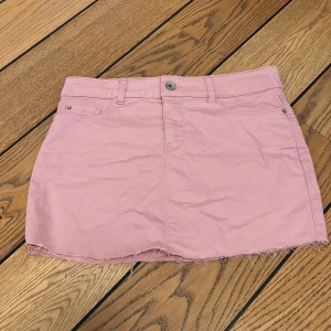 Rosa jeanskjol - Säljer en söt rosa jeanskjol med rå kant och klassisk femficksdesign. Kjolen har en knapp och dragkedja framtill samt hällor för bälte. Perfekt för en avslappnad stil!