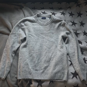 Grå ulltröja från GANT - Säljer en stilren grå ulltröja från GANT med rund hals och ribbade muddar. Perfekt för kyliga dagar och enkel att matcha med olika outfits. Tröjan har en diskret logga på bröstet. Perfekt att ha över en skjorta✅