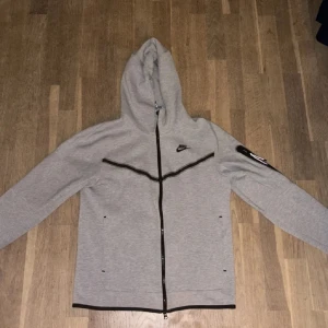 Grå Nike tech  - Säljer en grå hoodie från Nike med dragkedja och huva. Den har svarta detaljer och en ficka på ärmen. Perfekt för en avslappnad stil.