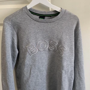 Grå tröja från Boss - Säljer en stilren grå tröja från Boss med broderad logga på bröstet. Tröjan har långa ärmar och en rund halsringning. Perfekt för en avslappnad look.