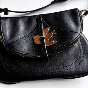 Svart axelväska från Marc Jacobs - Underbar svart axelväska från Marc Jacobs i skinn med en unik guldfärgad fågeldetalj på framsidan. Väskan har en justerbar axelrem (max längd ca 97 cm ) och stängs med magnetlås. Insidan är fodrad med det ikoniska tyget. Nyskick.