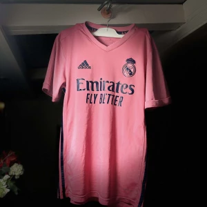 Rosa fotbollströja - Real Madrid - Säljer en snygg rosa fotbollströja från Real Madrid med korta ärmar. Tröjan har en V-ringning och klubbmärke på bröstet. Den har även texten 'Emirates Fly Better' tryckt på framsidan. Perfekt för fotbollsfans som vill sticka ut på planen!