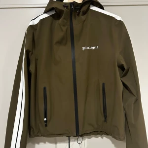 Olivgrön jacka från Palm Angels - Säljer min killes olivgröna jacka från Palm Angels med vita detaljer och dragkedja. Jackan har långa ärmar med vita ränder längs sidorna och en huva. Perfekt för en stilren look. Nypris 7000