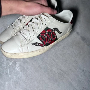 Vita sneakers med ormmotiv - Snygga vita sneakers med ett coolt ormmotiv på sidan. Skorna har snörning och en rutig detalj i rött och grönt. Perfekta för att ge din outfit en unik touch.