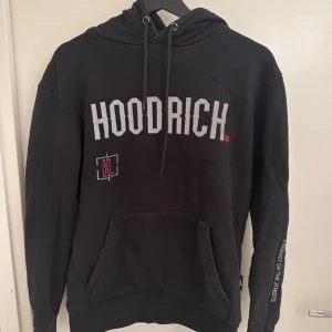 Svart hoodie från Hoodrich - Säljer en svart hoodie från Hoodrich med vit text på bröstet och en liten röd detalj. Den har en känguruficka och justerbar huva med dragsko. Perfekt för en avslappnad stil.