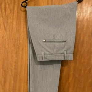 Grå kostymbyxor från Tailored Originals - Snygga grå kostymbyxor från Tailored Originals med en stilren design. Byxorna har en normal passform och är tillverkade i ett lätt och bekvämt material. De har knappar och dragkedja framtill samt eleganta bakfickor med knappar. Hittar ingen storlek men runt M/L tror jag. Byxorna liknar dem som är på dem sista två bilderna