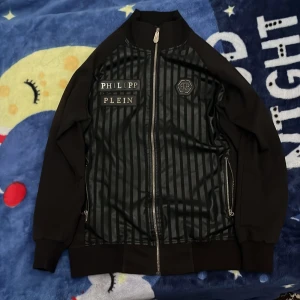 Svart jacka från Philipp Plein - Snygg svart jacka från Philipp Plein med dragkedja framtill. Framsidan har ett randigt mönster och märkeslogga, medan baksidan pryds av en stor dödskalle och röd text. Perfekt för en cool och edgy stil. M men passar till L också 