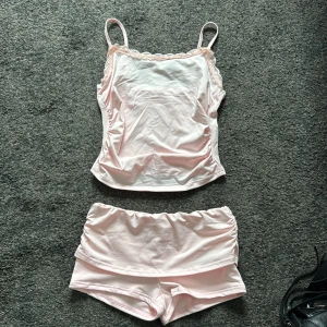 Ljusrosa linne och shorts set - Säljer ett sött ljusrosa set med linne och shorts. Linnet har smala axelband och spetsdetaljer vid halsen. Perfekt för en avslappnad dag hemma eller som pyjamas. Helt oanvänd!