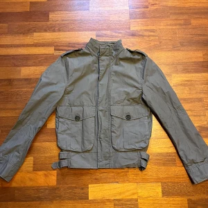 Grön overshirt Tiger of Sweden - Säljer en stilren grön jacka från Tiger of Sweden. Jackan har långa ärmar och praktiska fickor med knappar framtill. Den är tillverkad i ett syntetmaterial och har justerbara remmar i midjan för en bättre passform.