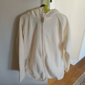 Vit fleecejacka med dragkedja - Mysig vit fleecejacka med dragkedja och huva. Perfekt för kyliga dagar. Jackan har två fickor med dragkedja framtill.
