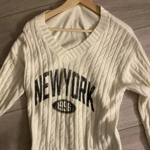 Vit stickad tröja med New York-tryck - Säljer en vit stickad tröja med ett coolt New York-tryck och årtalet 1996 på framsidan. Tröjan har en v-ringad hals och långa ärmar, perfekt för en avslappnad stil. strl S