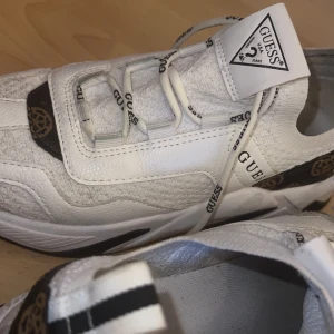 Vita sneakers från Guess - Snygga vita sneakers från Guess med svarta detaljer och logotyp på sidan. Skorna har snörning och en modern design med en bekväm sula. Perfekta för en stilren look.