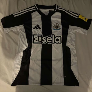 Newcastle United hemma 24/25 fotbollströja  - Säljer en svartvit randig fotbollströja från Adidas med korta ärmar. Tröjan har klubbmärke på bröstet och sponsorlogga på framsidan. Perfekt för fotbollsfans som vill visa sitt stöd.