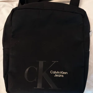 Svart axelväska från Calvin Klein Jeans - Snygg svart axelväska från Calvin Klein Jeans med logotypen tryckt på framsidan. Väskan har en praktisk dragkedja och justerbar axelrem. Perfekt för att bära dina dagliga nödvändigheter med stil.