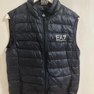 Svart dunväst från Emporio Armani - Snygg svart dunväst från Emporio Armani med dragkedja framtill. Västen har en stilren design med märkets logga på bröstet. Perfekt för lager-på-lager under kyligare dagar.