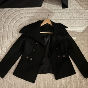 Svart kappa från Gina Tricot - Snygg svart kappa från Gina Tricot med stora knappar och en elegant krage. Jackan har en figursydd passform och är perfekt för kyligare dagar. Den har långa ärmar och ett stilrent bälte i midjan för extra stilpoäng. Säljer då den är för liten för mig 