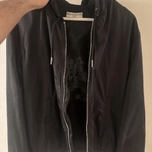 Svart vindjacka från Givenchy - Snygg svart vindjacka från Givenchy med blommigt mönster och logotyp på ryggen. Jackan har dragkedja framtill och en huva. Ny pris för denna jackan är ungefär 15 000kr. Passar inte mig längre då jag gått upp i vikt en del.