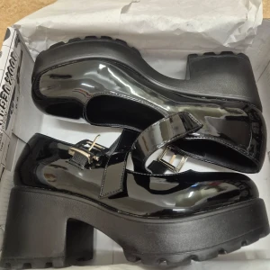 Koi Footwear Tira Mary Jane 37 - Säljer ett par svarta Mary Jane skor i lack med bred platåsula och klack. Skorna har ett justerbart spänne i silver och rund tå. Använd en gång. I princip nyskick. 