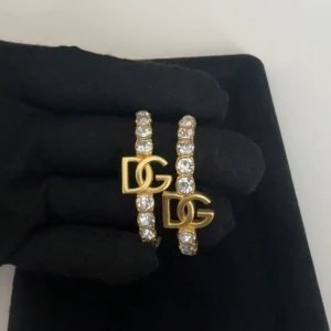 Guldiga DG örhängen med stenar - Säljer ett par guldiga örhängen med DG-logga. Örhängena är halvcirkelformade och dekorerade med gnistrande stenar längs hela bågen. Perfekta för att ge din look extra lyxig känsla.
