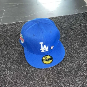 Blå LA Dodgers keps New Era 59FIFTY - Säljer en blå LA Dodgers keps från New Era 59FIFTY med broderad LA-logga framtill, 1988 World Series-märke på sidan och texten 'Love & Hustle is the Game' bak. Klassisk flat peak och MLB-logga bak.