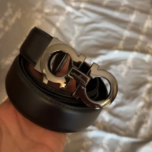 Svart skärp med silverfärgat spänne från Salvatore Ferragamo - Snyggt svart bälte i skinn med ett stort, blankt silverfärgat spänne i Ferragamos ikoniska design. Perfekt accessoar för att lyfta din outfit med en lyxig touch.