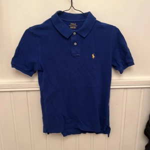 Blå piké från Polo Ralph Lauren - Klassisk blå pikétröja från Polo Ralph Lauren med korta ärmar och krage. Tröjan har två knappar vid halsen och den ikoniska gula broderade loggan på bröstet. Perfekt för en stilren och sportig look.