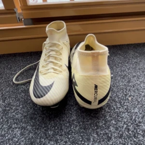 Vita Nike Mercurial fotbollsskor med svart swoosh - Säljer ett par vita Nike Mercurial fotbollsskor med svart swoosh och snörning. Skorna har hög krage och dobbar för bra grepp på planen. Perfekta för dig som vill ha snabba och lätta skor till match eller träning.
