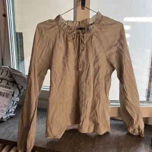 Beige blus med volangkrage från Vero Moda - Säljer en beige blus från Vero Moda med lång ärm och volangdetalj vid kragen. Blusen har knytning framtill och resår vid ärmsluten för en snygg passform. Perfekt för dig som gillar stilrena och feminina plagg.